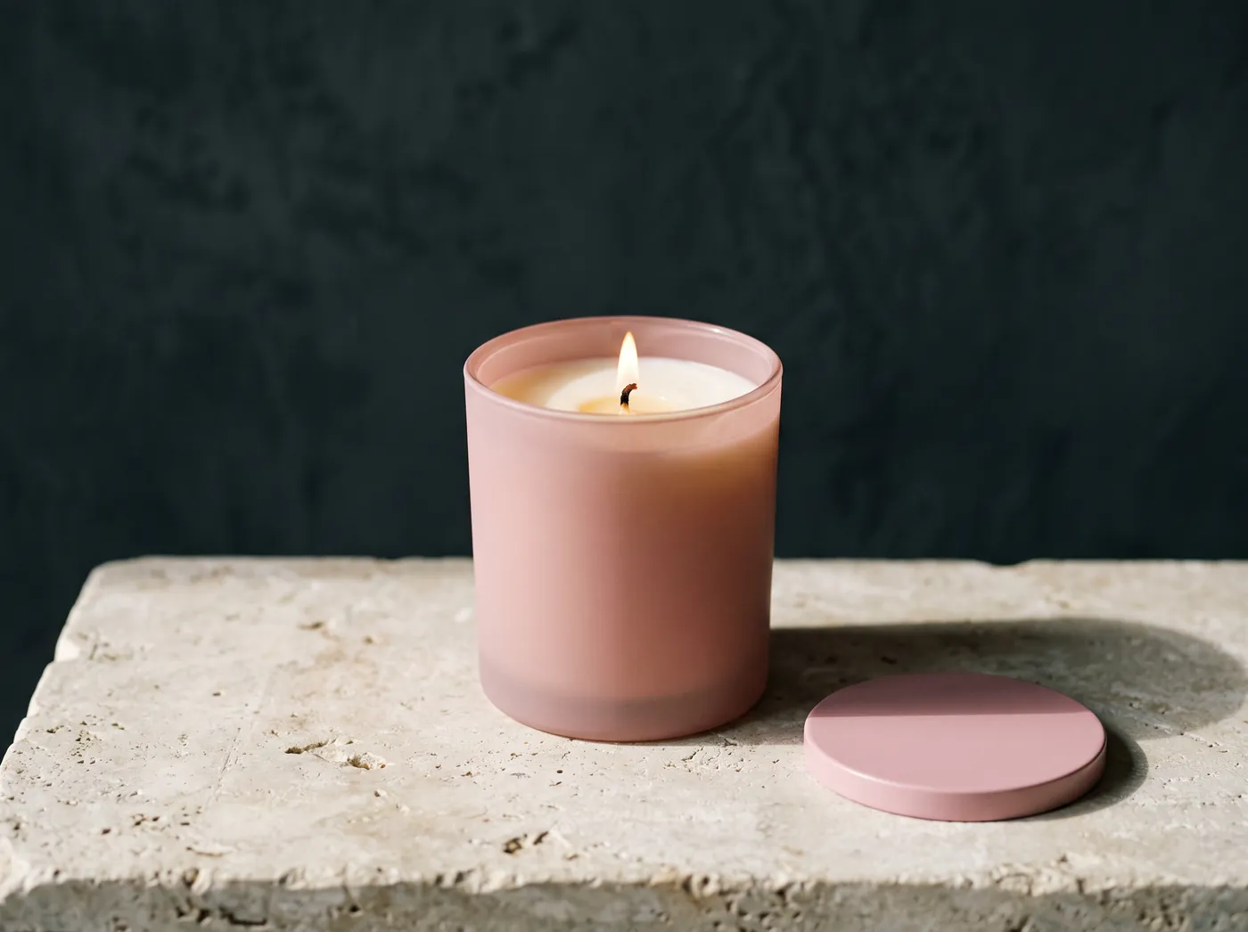 A lit candle in a matte dusty pink frosted glass tumbler with its flat pink lid resting on the slab beside it. Vessel study, not a product photograph.