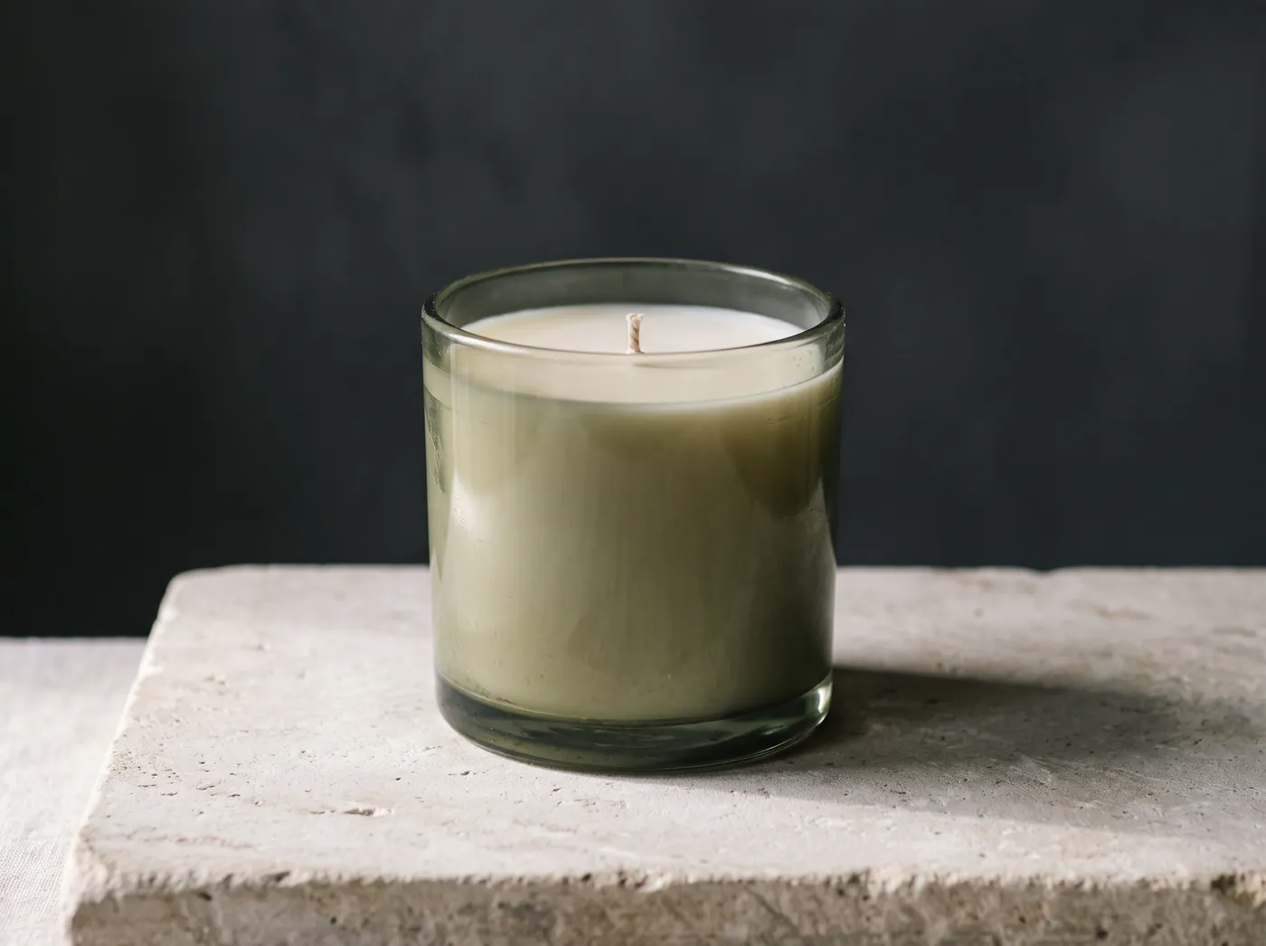 An unlit candle in a heavy sage-olive coloured glass vessel with thick walls and a flat pristine wax surface. Vessel study, not a product photograph.