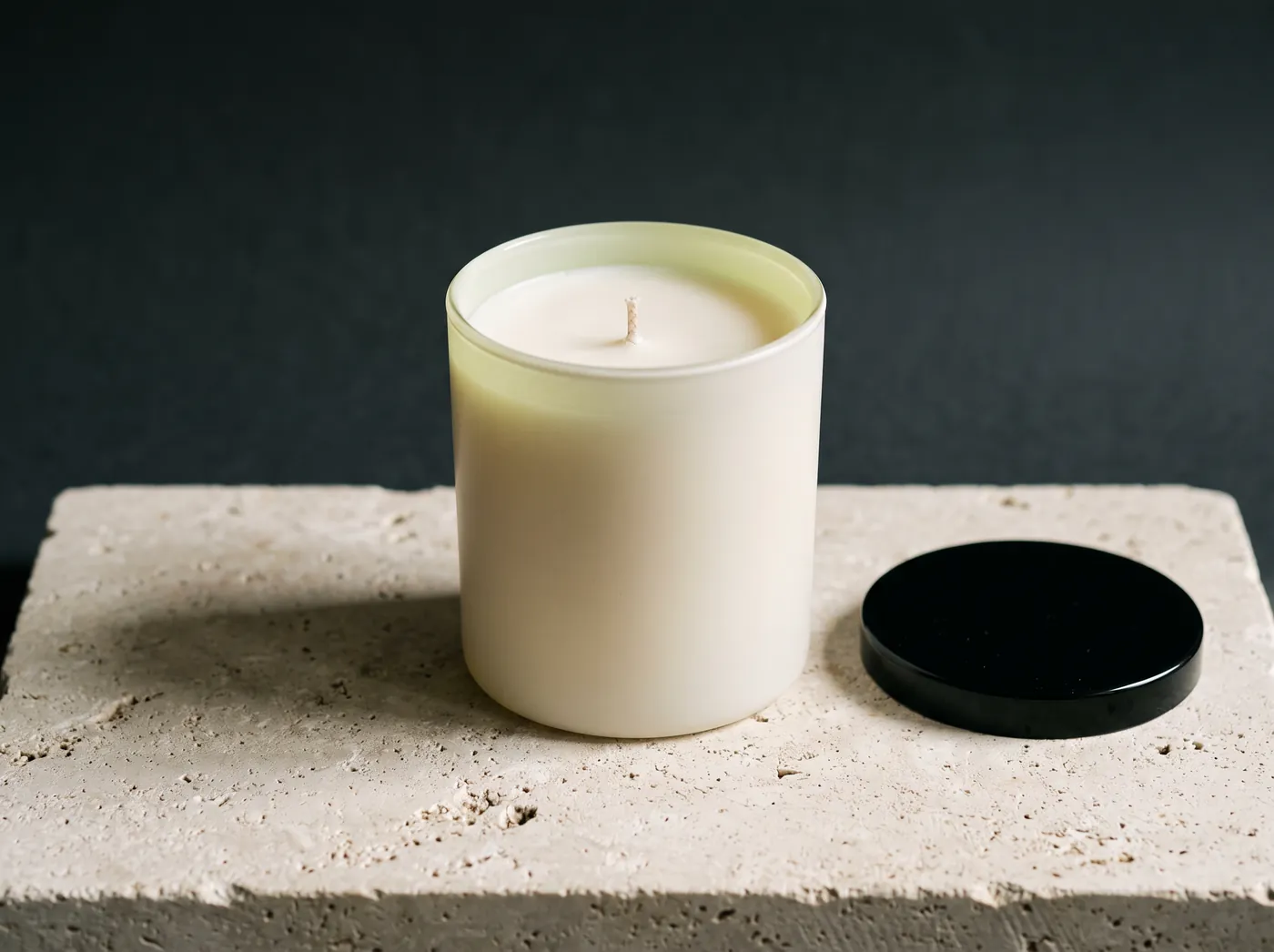 An unlit candle in an opaque cream glass jar with a black lacquered lid lying flat on the slab beside it. Vessel study, not a product photograph.