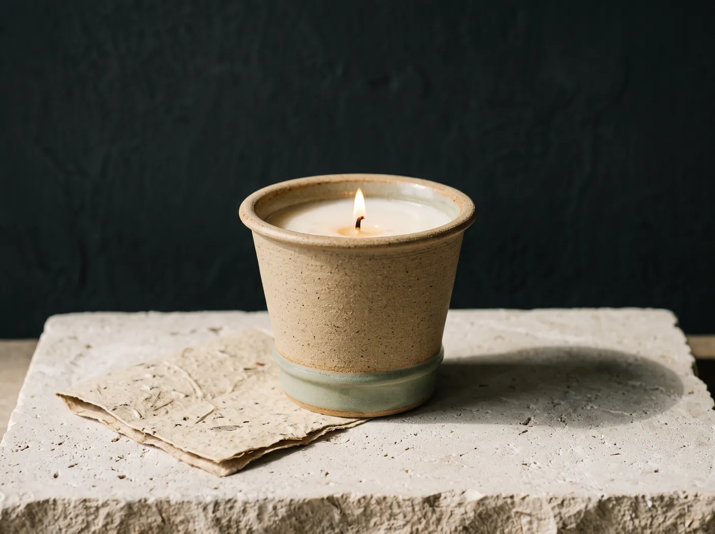 A lit candle in a tapered oatmeal stoneware planter with a sage green glaze band at the foot, on a limestone slab beside a folded sheet of seed paper. Vessel study, not a product photograph.
