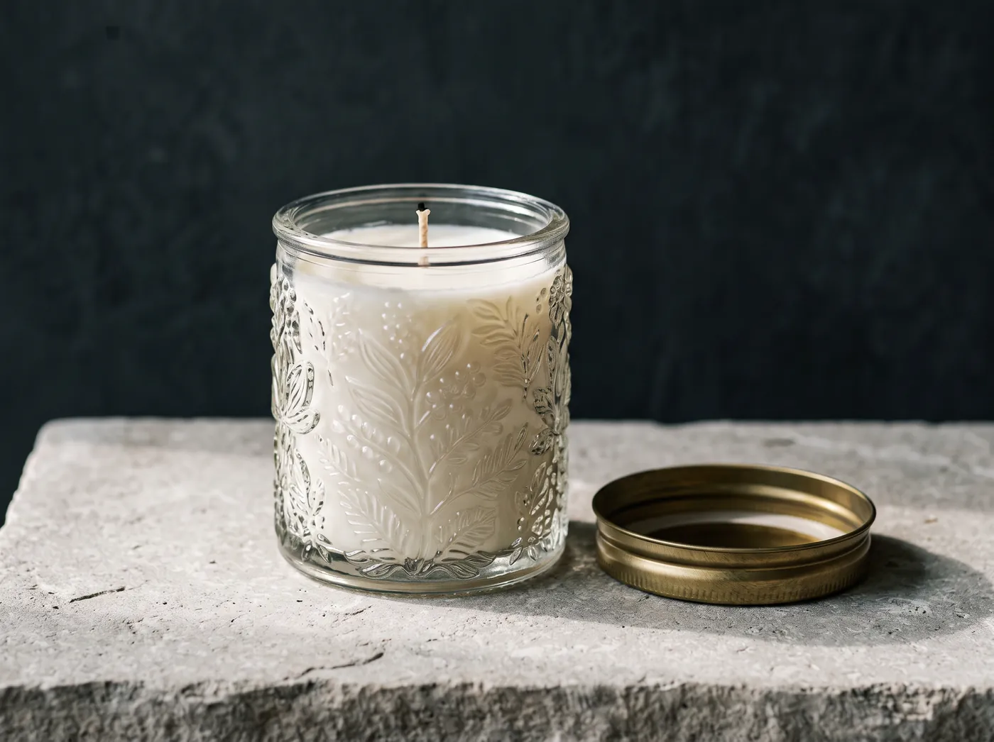 An unlit candle in an ornate embossed glass jar with a raised botanical relief, its brushed gold lid on the slab beside it. Vessel study, not a product photograph.
