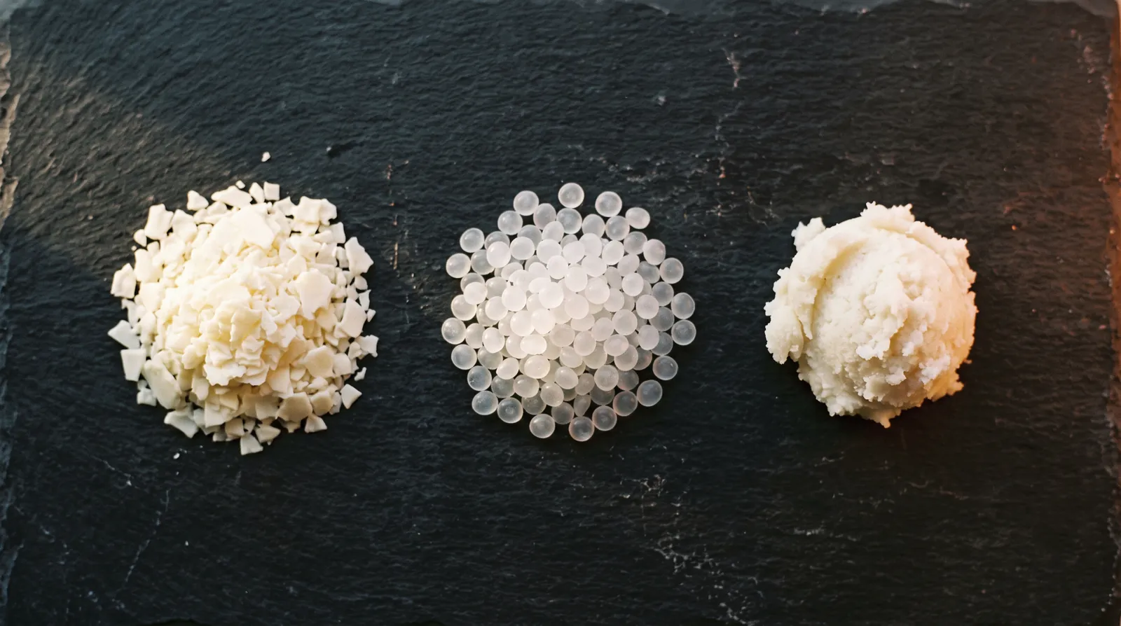 Soy wax flakes, translucent paraffin pellets and soft coconut wax in three piles on a slate slab